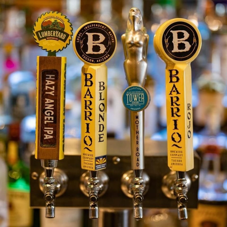 Four craft beer taps display Lumberyard, Barrio, Blonde, Mother Road, and Roja labels against a softly blurred bar background.