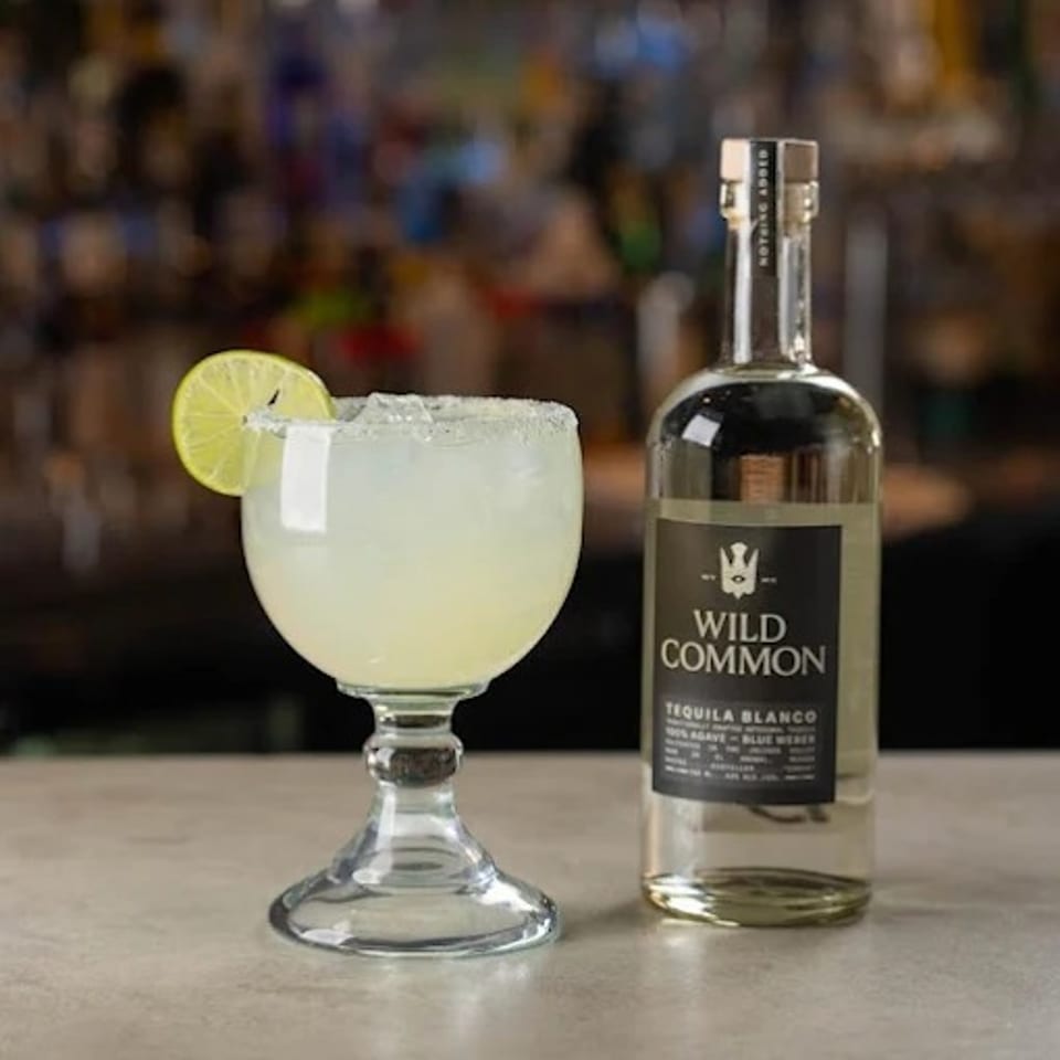 Margarita with lime garnish beside a bottle of Wild Common tequila on a bar counter.