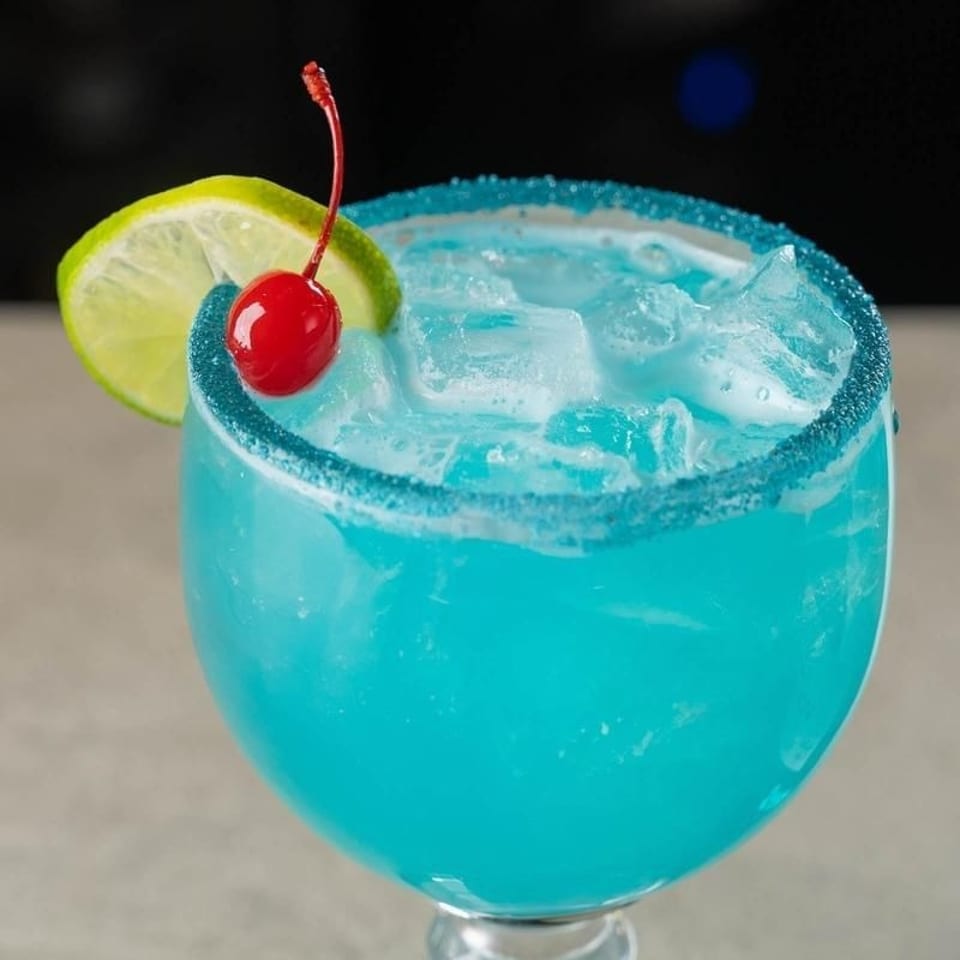 Bright blue cocktail with ice, a sugar-rimmed glass, lime slice, and red cherry garnish.