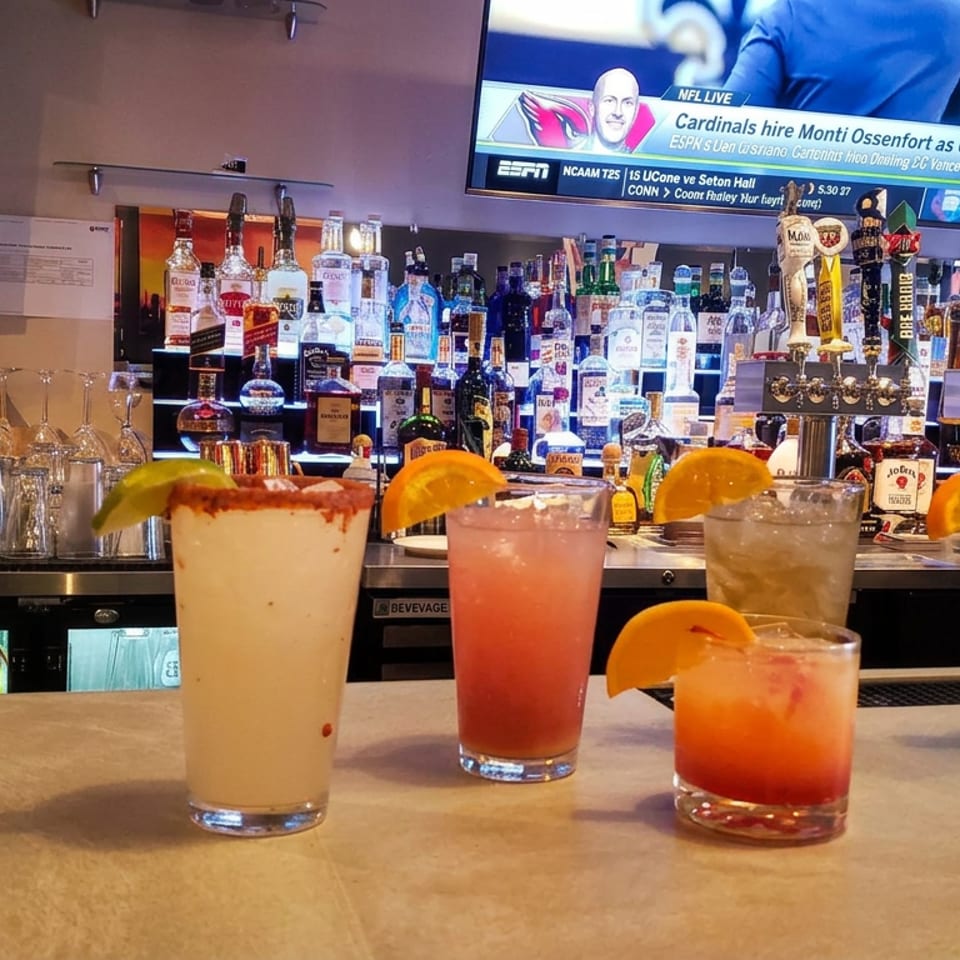 Five colorful cocktails garnished with orange wedges sit on a bar, backed by liquor bottles, beer taps, and sports televisions.