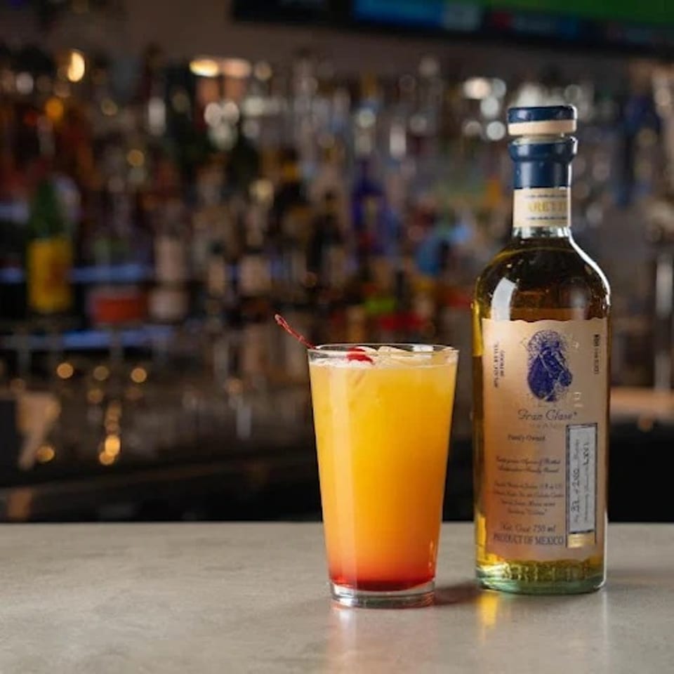 Orange-red cocktail with citrus garnish beside a bottle of tequila on a bar counter, with a blurred liquor shelf in the background.