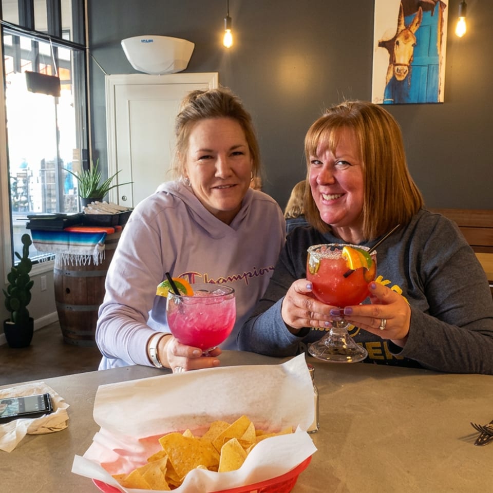 Two women smile while holding colorful cocktails at a restaurant table, with tortilla chips, water, and warm pendant lighting visible.