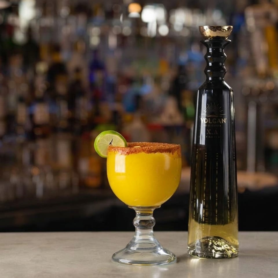 Bright yellow cocktail with a chili-salt rim and lime garnish beside a Volcan tequila bottle on a bar.