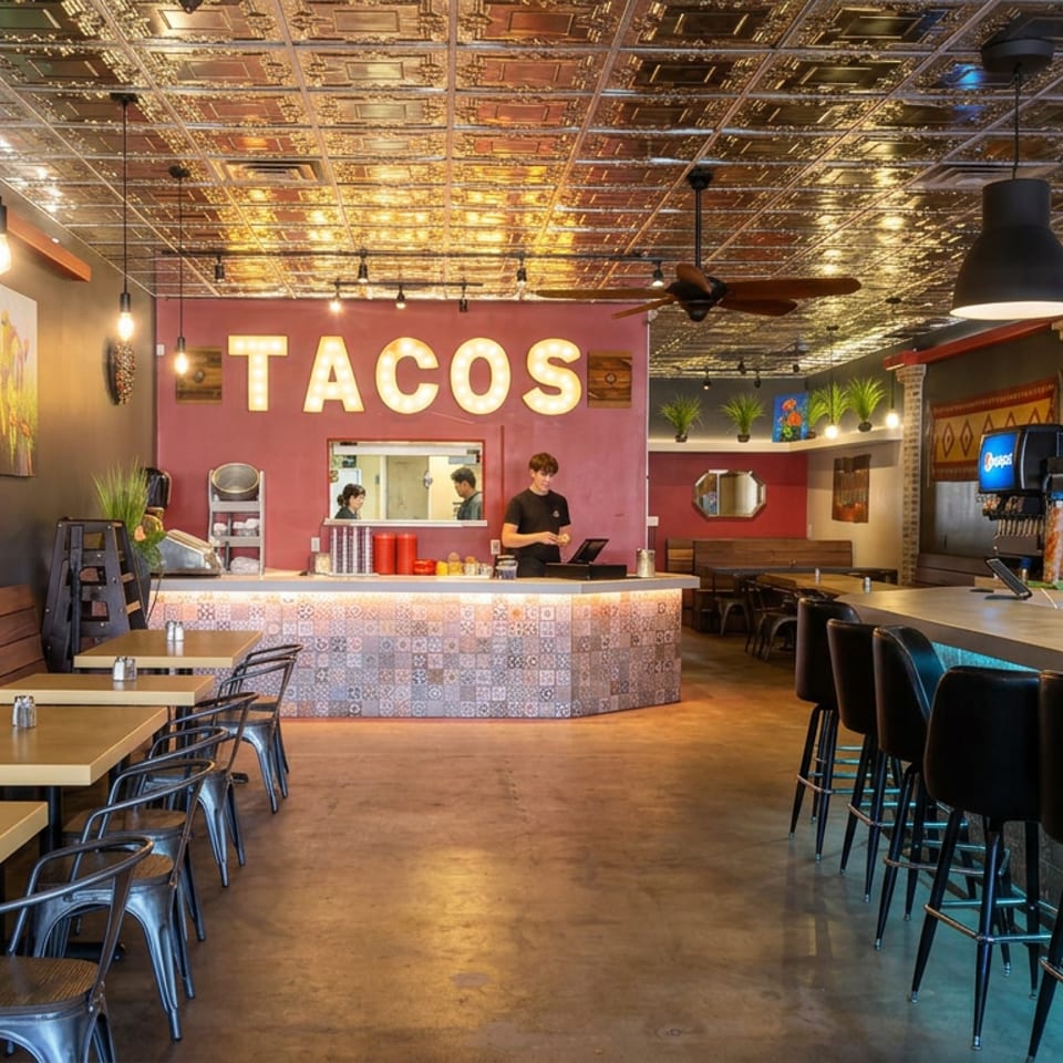 Colorful taco restaurant interior with patterned ceiling, illuminated taco sign, counter seating, tables, and staff behind the counter.