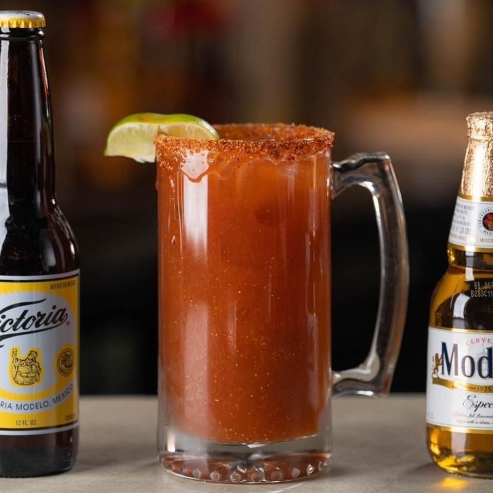 Spicy red beer cocktail in a salt-rimmed mug with lime wedge, flanked by Victoria and Modelo beer bottles.