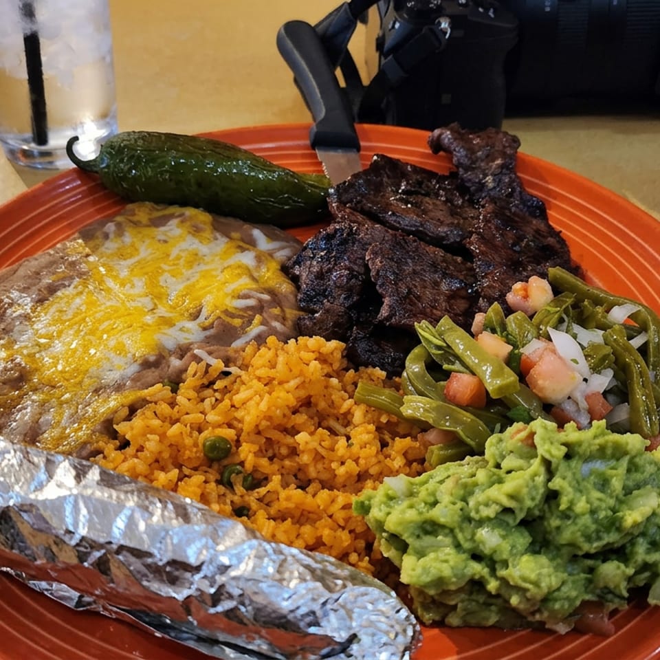 Mexican platter with grilled steak, cheesy refried beans, rice, guacamole, green beans, roasted pepper, and foil-wrapped corn.